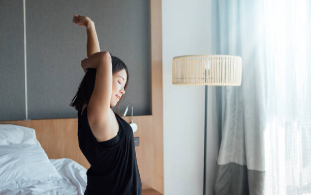 Side view of young Asian woman stretching arms after waking up in bed, with sunlight from the window in the morning.
